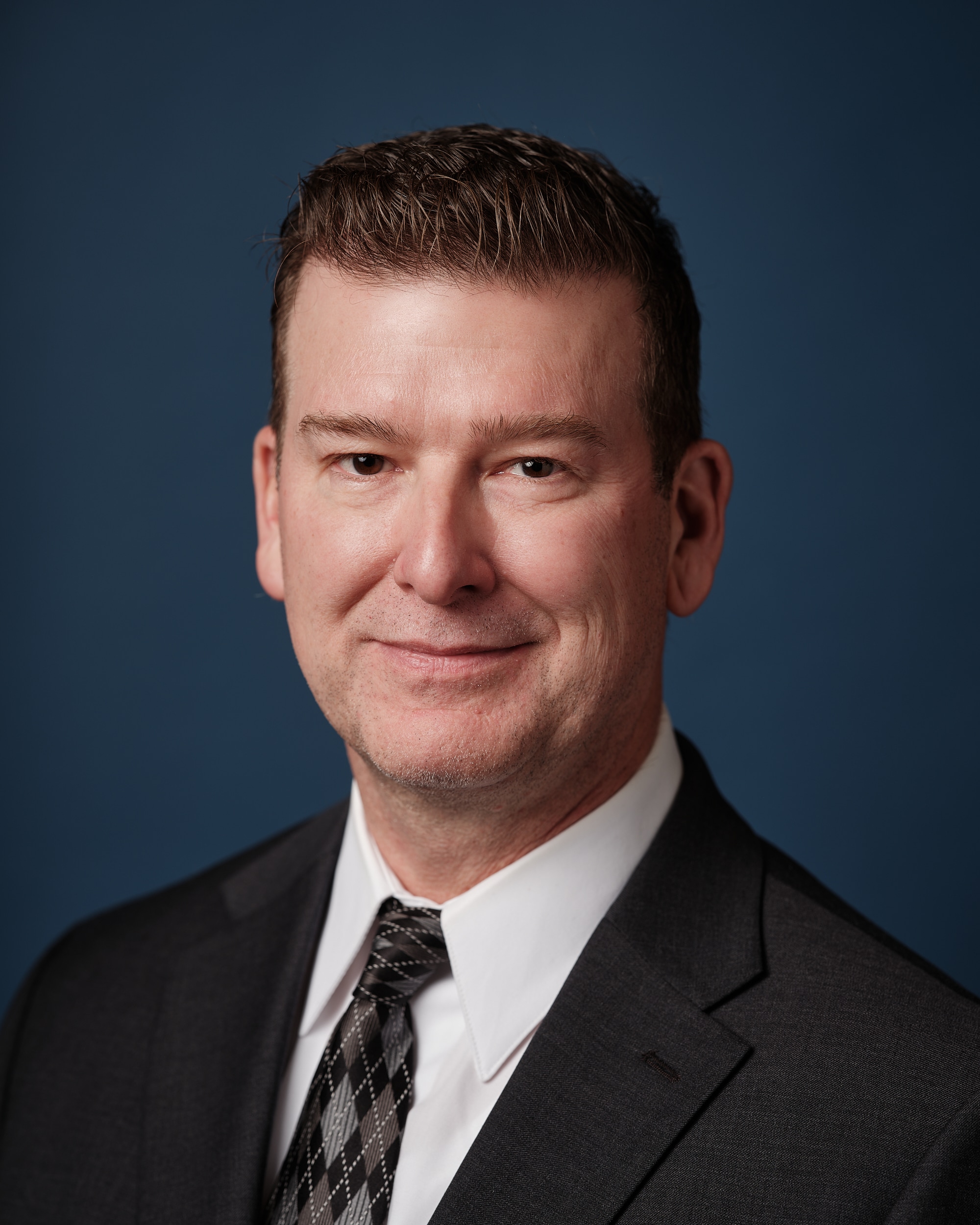 Head shot of Gary Waters with black patterned tie and dark grey suit with blue background.