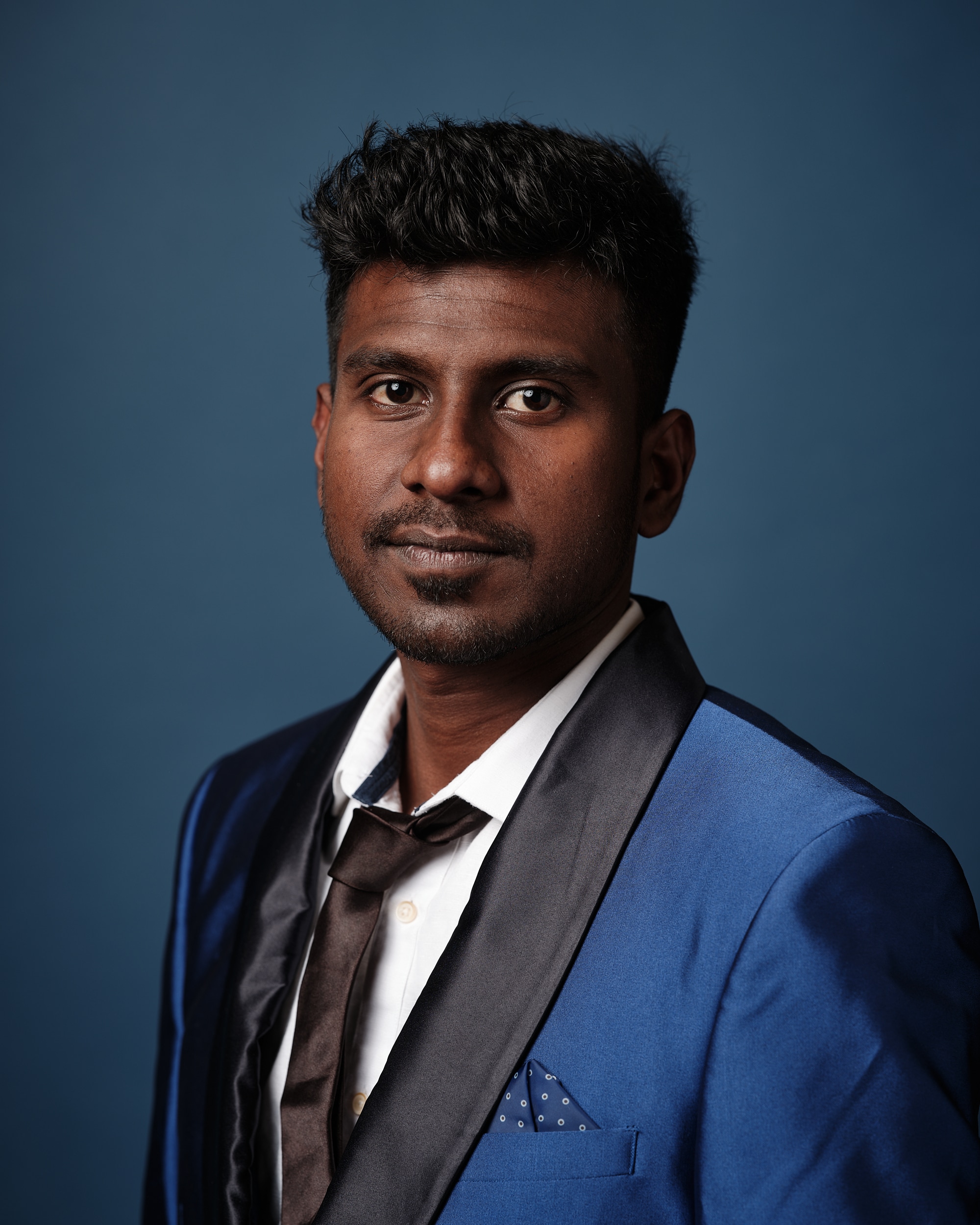 Professional headshot of a Jacob Thankamony in a blue suit jacket and tie, standing in front of a dark blue background.