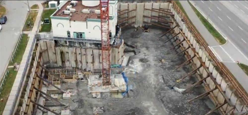 Aerial view of deep excavation site with concrete shoring and a crane at 935 Eglinton Avenue West, adjacent to a main road and existing structure.